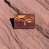 Moab Utah enamel pin with geometric orange and red mesas under a rising sun, lying on red sandstone in Moab, Utah.