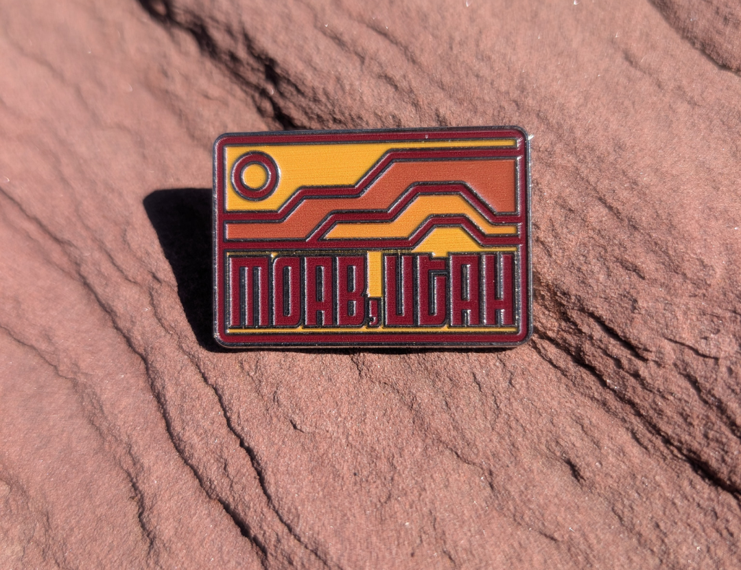 Moab Utah enamel pin with geometric orange and red mesas under a rising sun, lying on red sandstone in Moab, Utah.