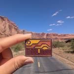 Hand holding the Moab Utah geometric sunset enamel pin on a road through the Moab red rock cliffs.