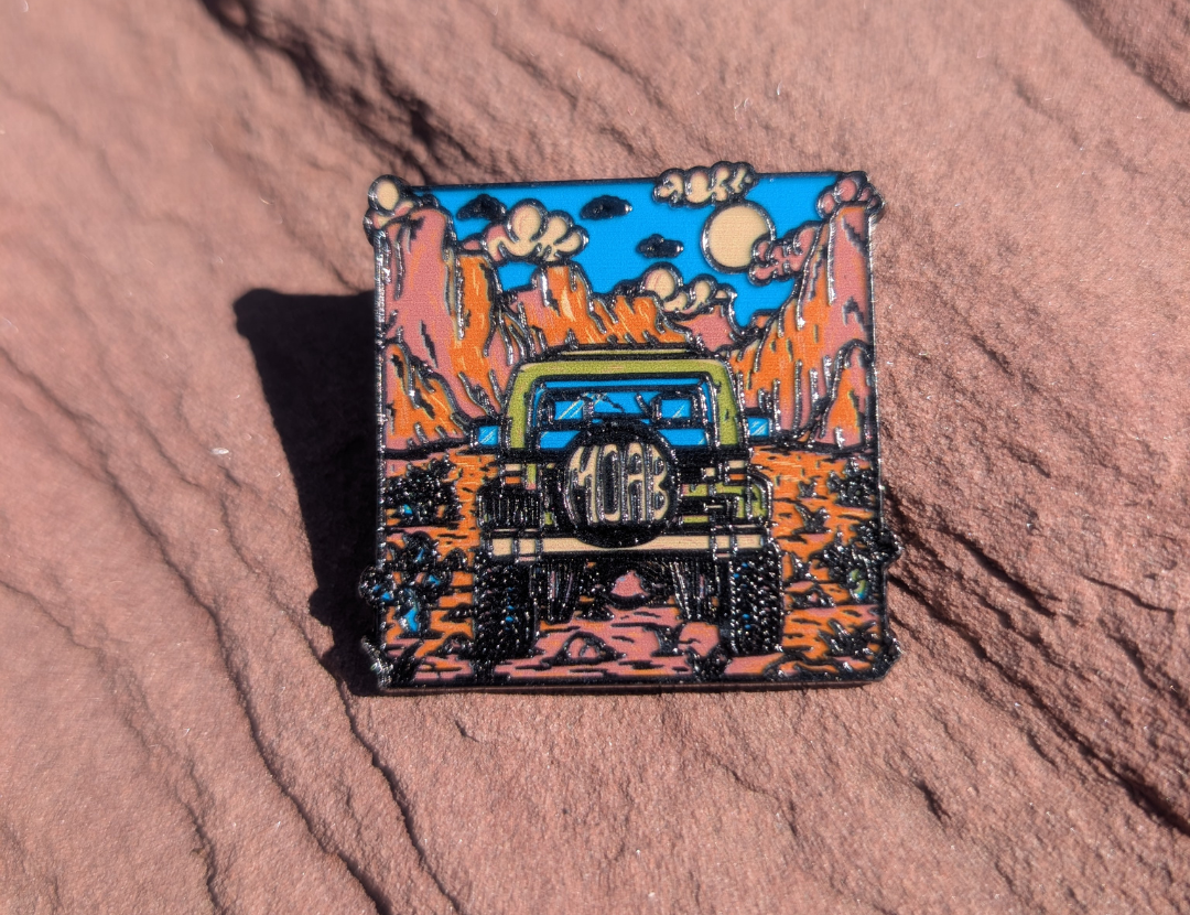 Square Moab enamel pin with a green Jeep climbing red rock under a blue sky, lying on red sandstone in Moab, Utah.