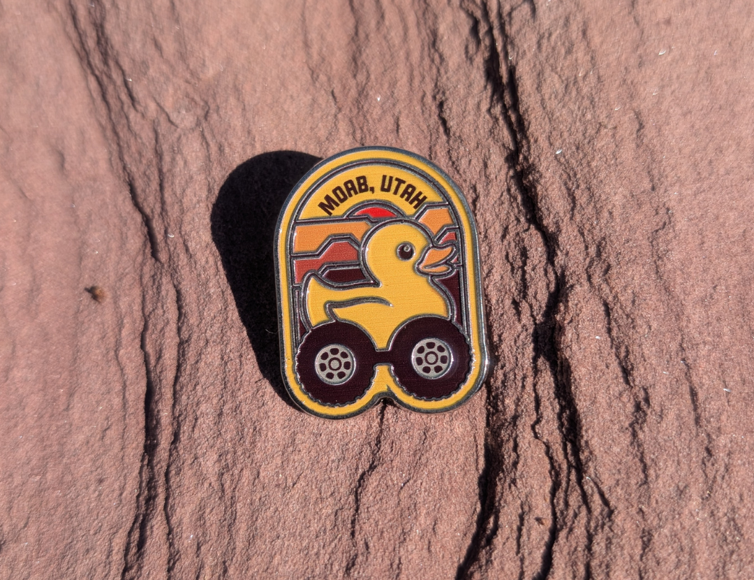 Arch shaped Moab Utah enamel pin with a yellow rubber duck riding on off road tires, lying on red sandstone in Moab, Utah.