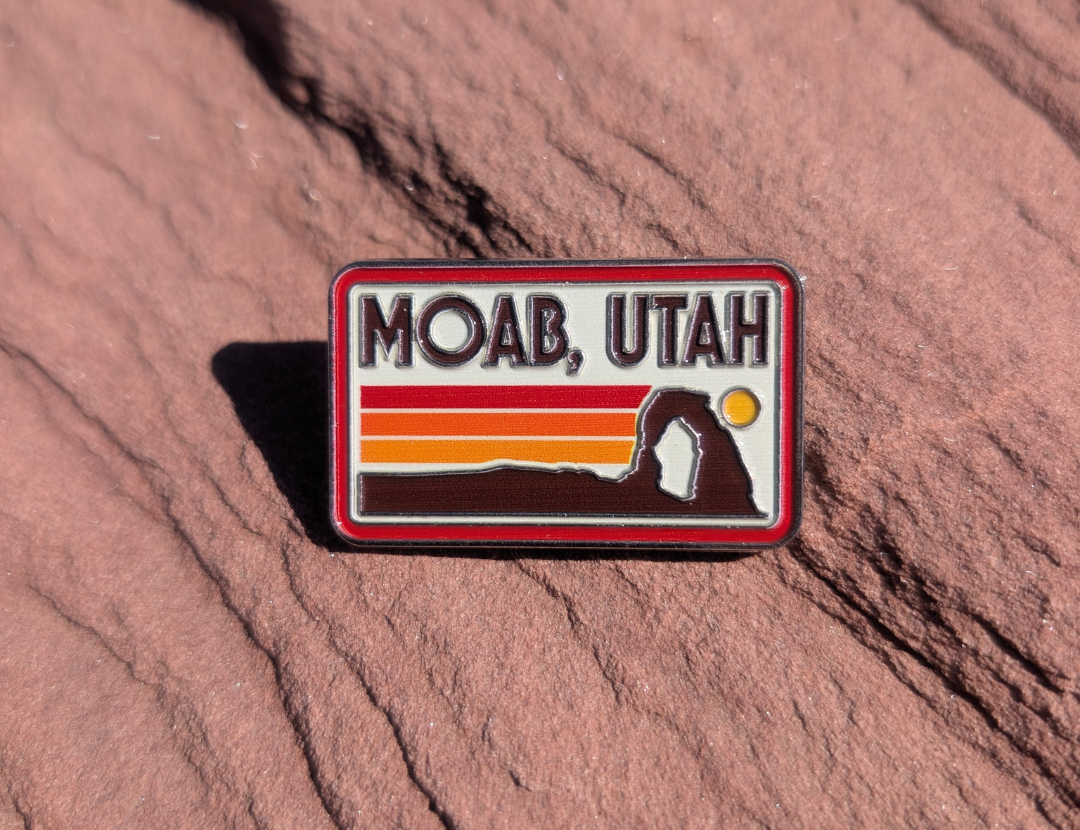 Rectangular Moab Utah enamel pin with retro sunset stripes and a Delicate Arch silhouette, lying on red sandstone in Moab, Utah.
