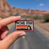 Hand holding the Moab Utah retro stripes enamel pin on a road through the Moab red rock cliffs.