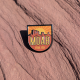Shield shaped Moab Utah USA enamel pin with a sunburst rising behind red mesas, lying on red sandstone in Moab, Utah.