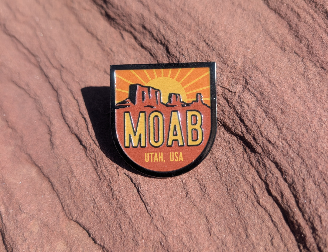 Shield shaped Moab Utah USA enamel pin with a sunburst rising behind red mesas, lying on red sandstone in Moab, Utah.
