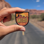 Hand holding the Moab Utah sunburst enamel pin on a road through the Moab red rock cliffs.