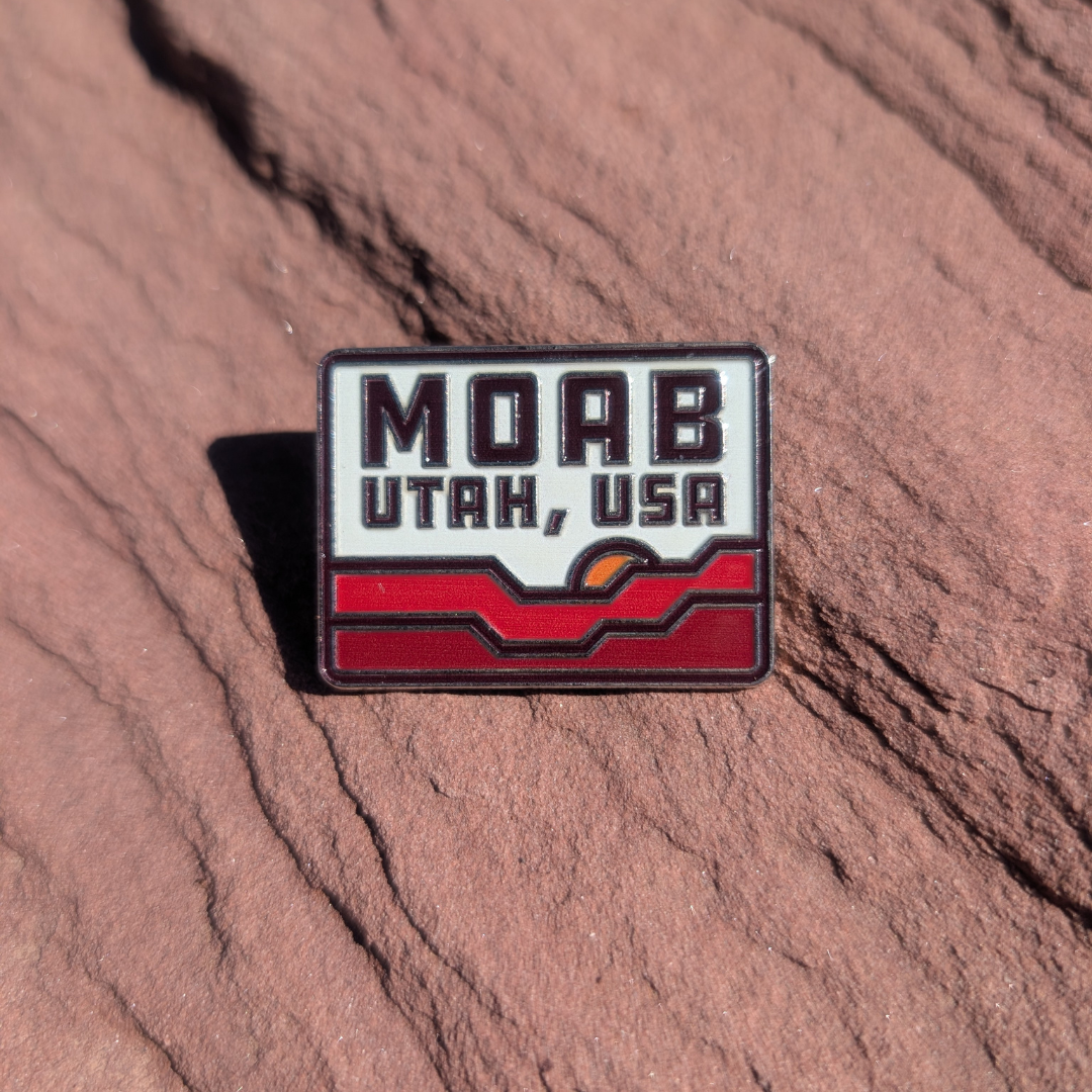 Rectangular Moab Utah USA enamel pin with red rock waves and a setting sun, lying on red sandstone in Moab, Utah.