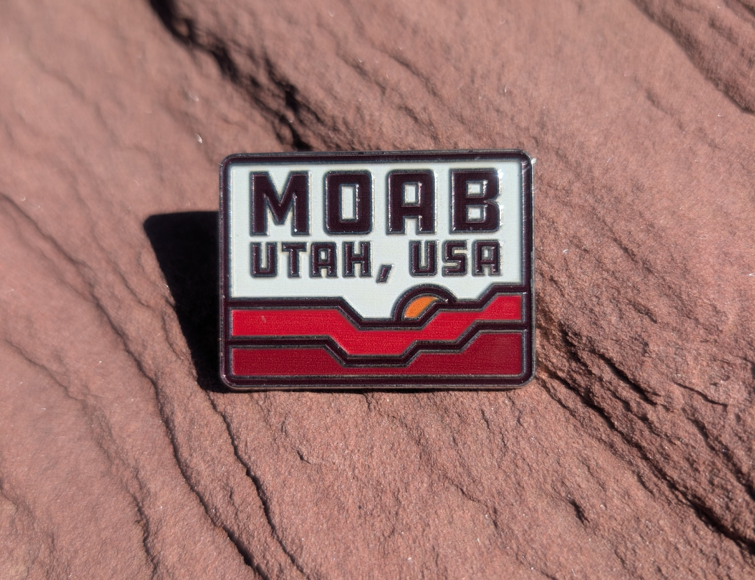 Rectangular Moab Utah USA enamel pin with red rock waves and a setting sun, lying on red sandstone in Moab, Utah.