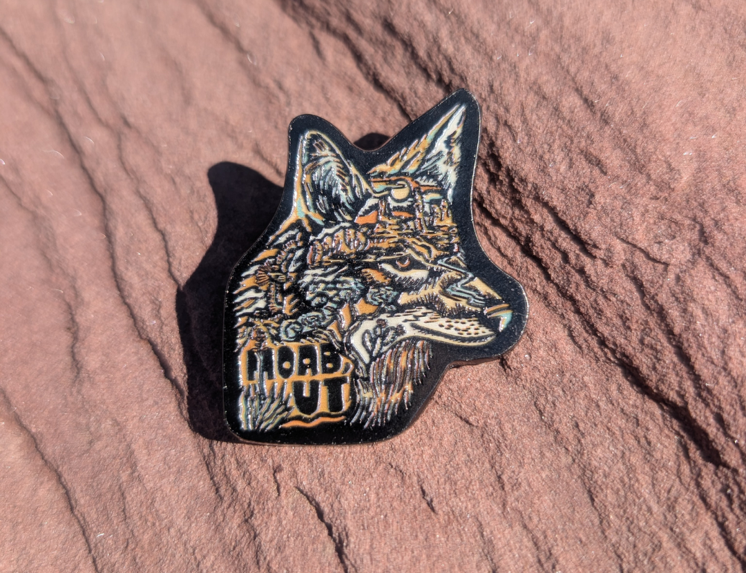 Die cut enamel pin of a wolf head in detailed line art with Moab UT lettering, lying on red sandstone in Moab, Utah.