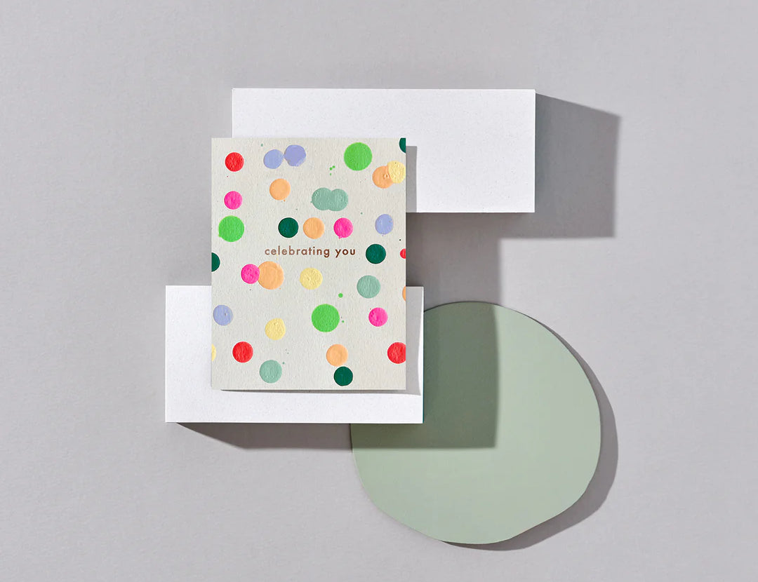 Moglea Celebrating Dot card styled on a gray surface with paper shapes and a sage green circle