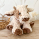 Mon Ami Caden Cow Plush sitting on a wood floor beside a wicker basket