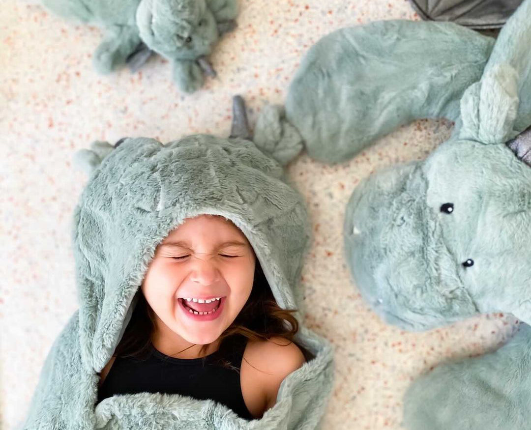 Laughing child in the Mon Ami Dax Dragon Plush Blanket hood lying among dragon plush toys