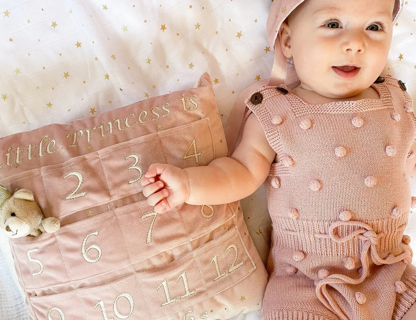 Baby in a pink outfit lying beside the Mon Ami My First Year Advent Pillow in Princess