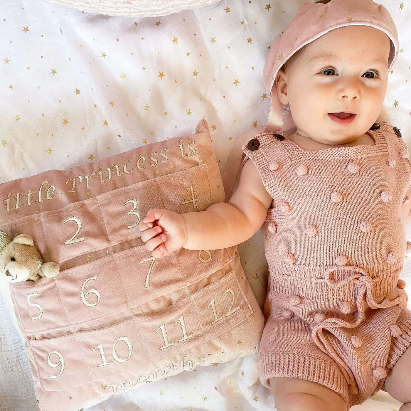 Baby in a pink outfit lying beside the Mon Ami My First Year Advent Pillow in Princess