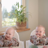 Two smiling babies in high chairs playing with Mushie Blossom Suction Spinner Toys by a sunny window