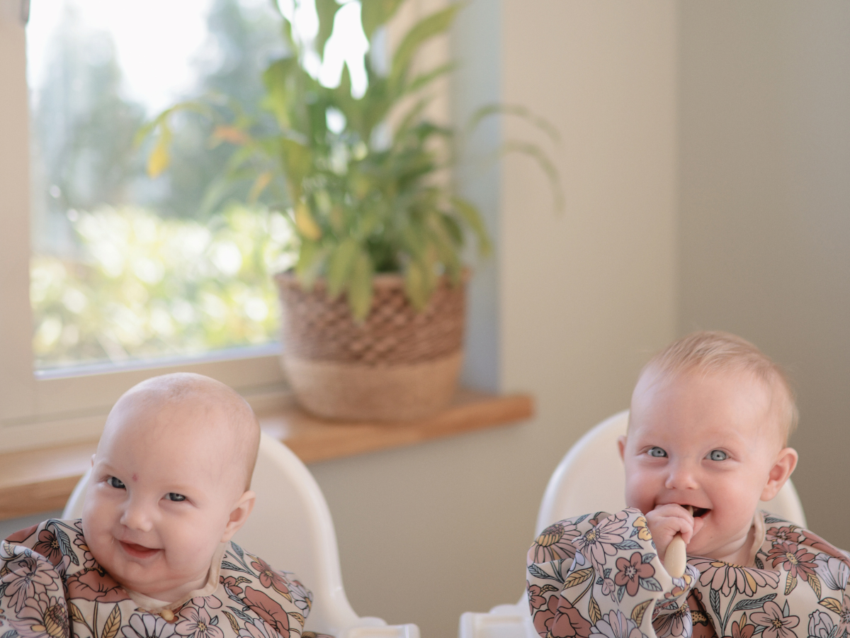 Two smiling babies in high chairs playing with Mushie Blossom Suction Spinner Toys by a sunny window