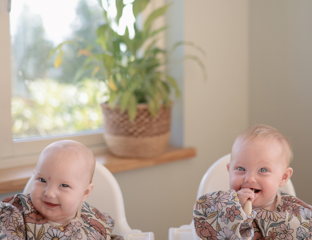 Two smiling babies in high chairs playing with Mushie Blossom Suction Spinner Toys by a sunny window