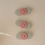 Three Mushie Blossom Suction Spinner Toys in sage green and pink lined up on a cream surface
