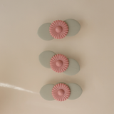 Three Mushie Blossom Suction Spinner Toys in sage green and pink lined up on a cream surface