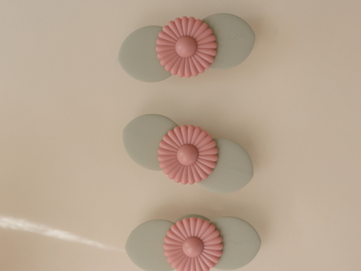Three Mushie Blossom Suction Spinner Toys in sage green and pink lined up on a cream surface