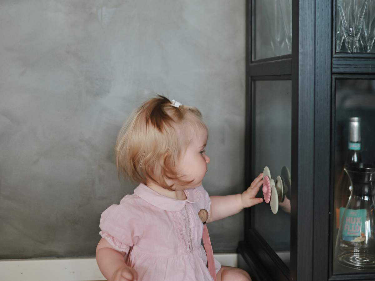Toddler in pink spinning a Mushie Blossom Suction Spinner Toy stuck to a glass cabinet door