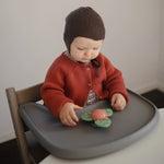 Baby in a red cardigan playing with the Mushie Dino Suction Spinner Toy on a high chair tray