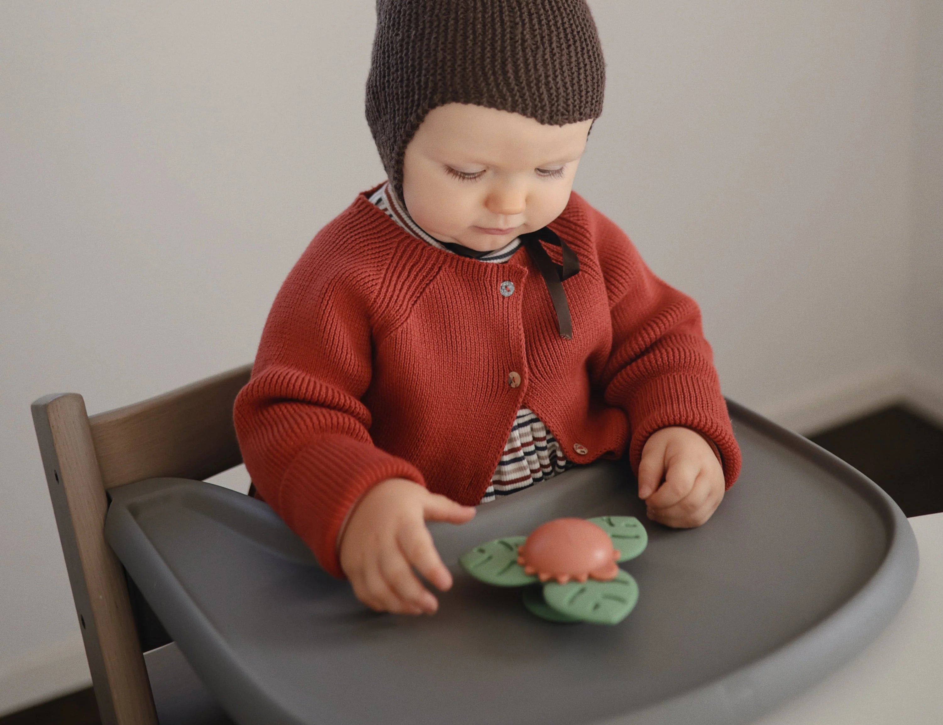 Baby in a red cardigan playing with the Mushie Dino Suction Spinner Toy on a high chair tray