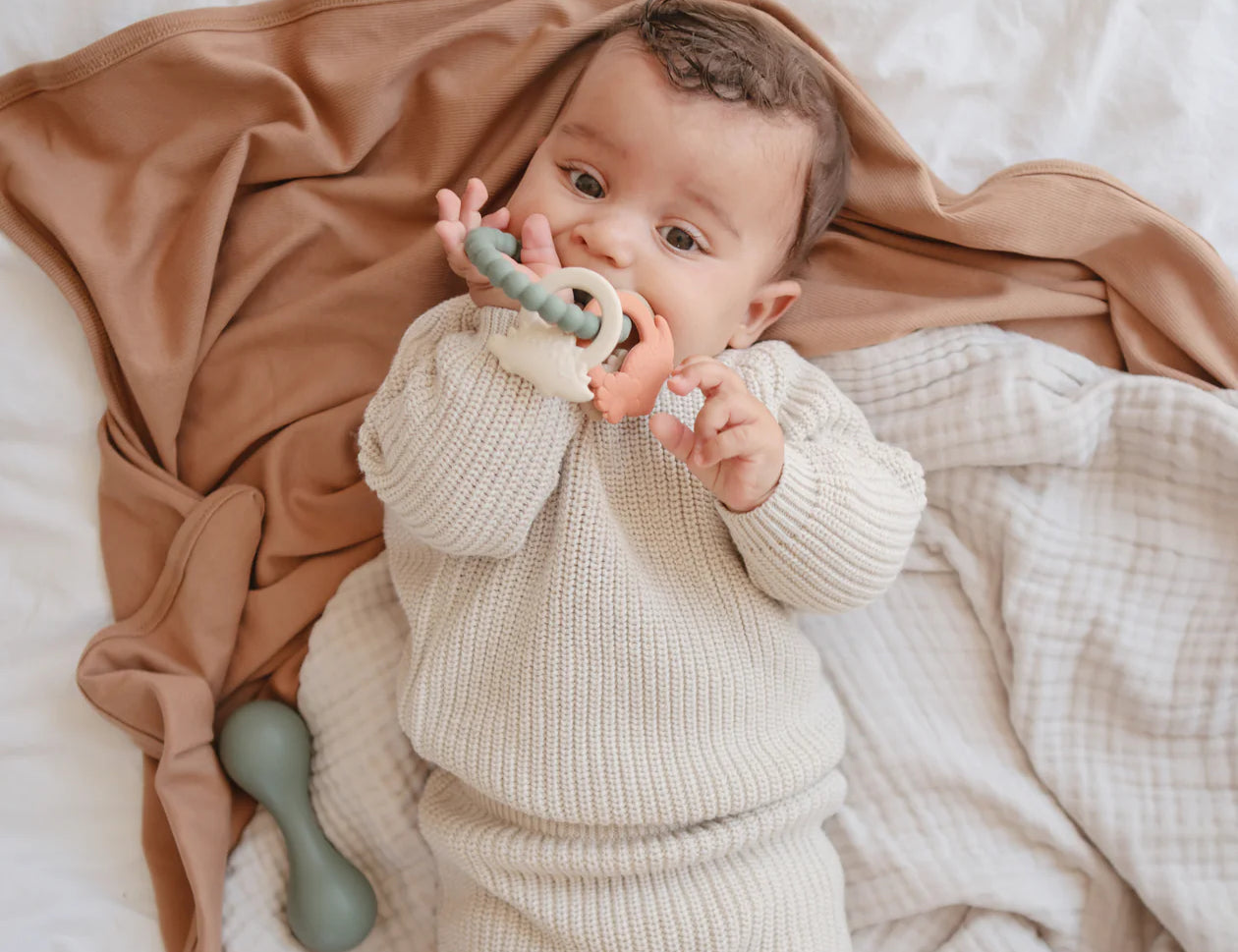 Baby lying on a blanket chewing on the Mushie Dino Teething Ring