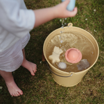 Child playing with the Mushie Mold Free Bath Play Set toys in a bucket of water on the grass