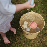 Child playing with the Mushie Mold Free Bath Play Set toys in a bucket of water on the grass