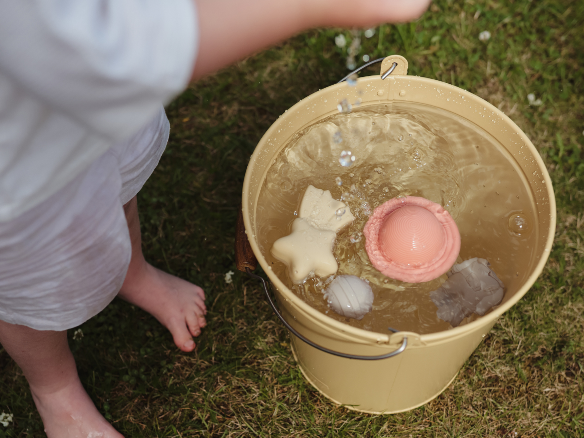 Child playing with the Mushie Mold Free Bath Play Set toys in a bucket of water on the grass
