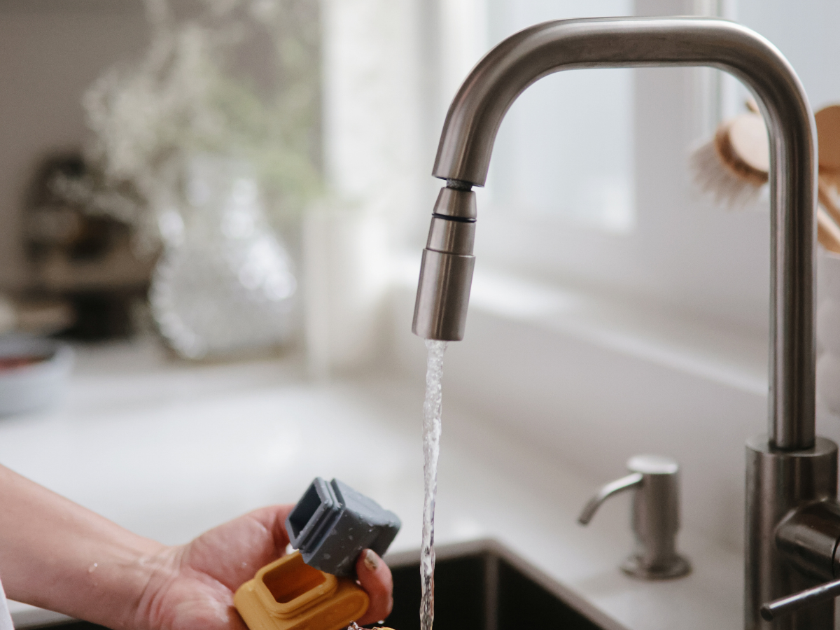 Hands rinsing a Mushie Mold Free Bath Play Set toy under a kitchen faucet