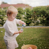 Toddler pouring water from a Mushie Mold Free Bath Play Set toy into a bucket in a garden