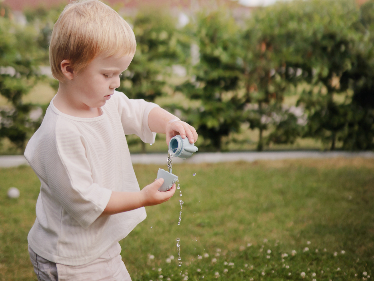 Toddler pouring water from a Mushie Mold Free Bath Play Set toy into a bucket in a garden