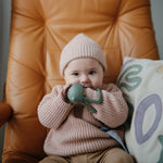Baby in a pink knit hat chewing the Mushie No-Drop Cactus Teether on a leather chair