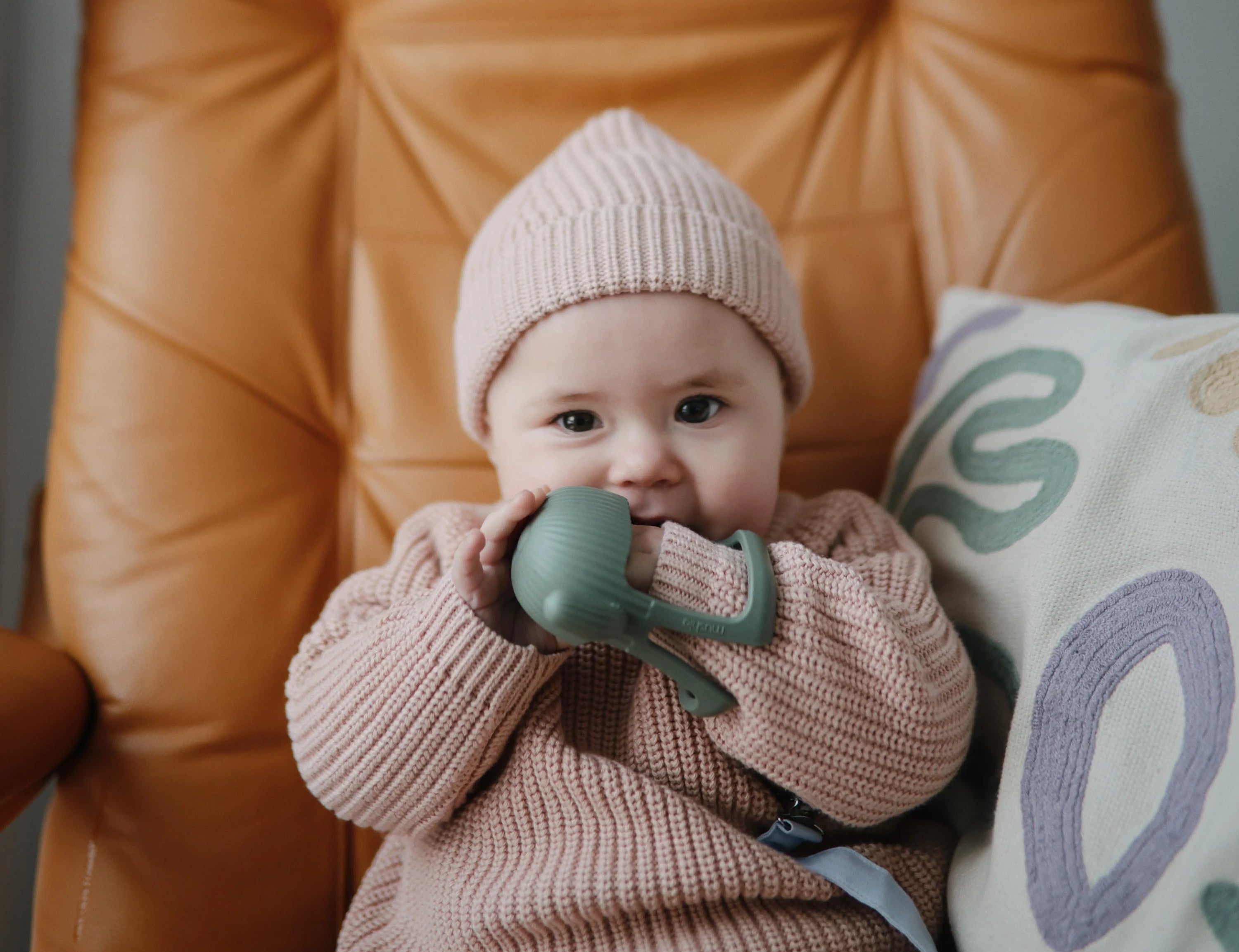 Baby in a pink knit hat chewing the Mushie No-Drop Cactus Teether on a leather chair