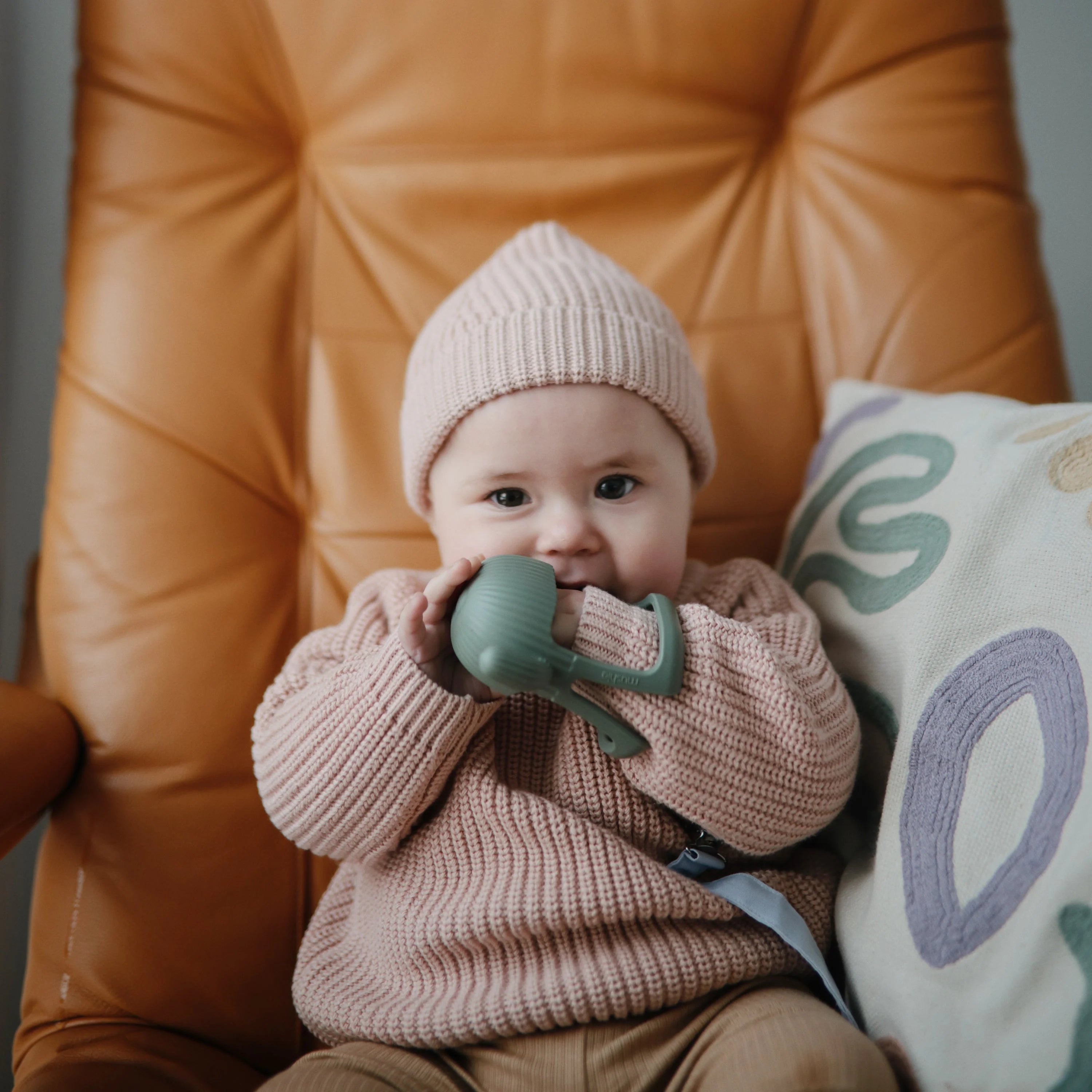 Baby in a pink knit hat chewing the Mushie No-Drop Cactus Teether on a leather chair
