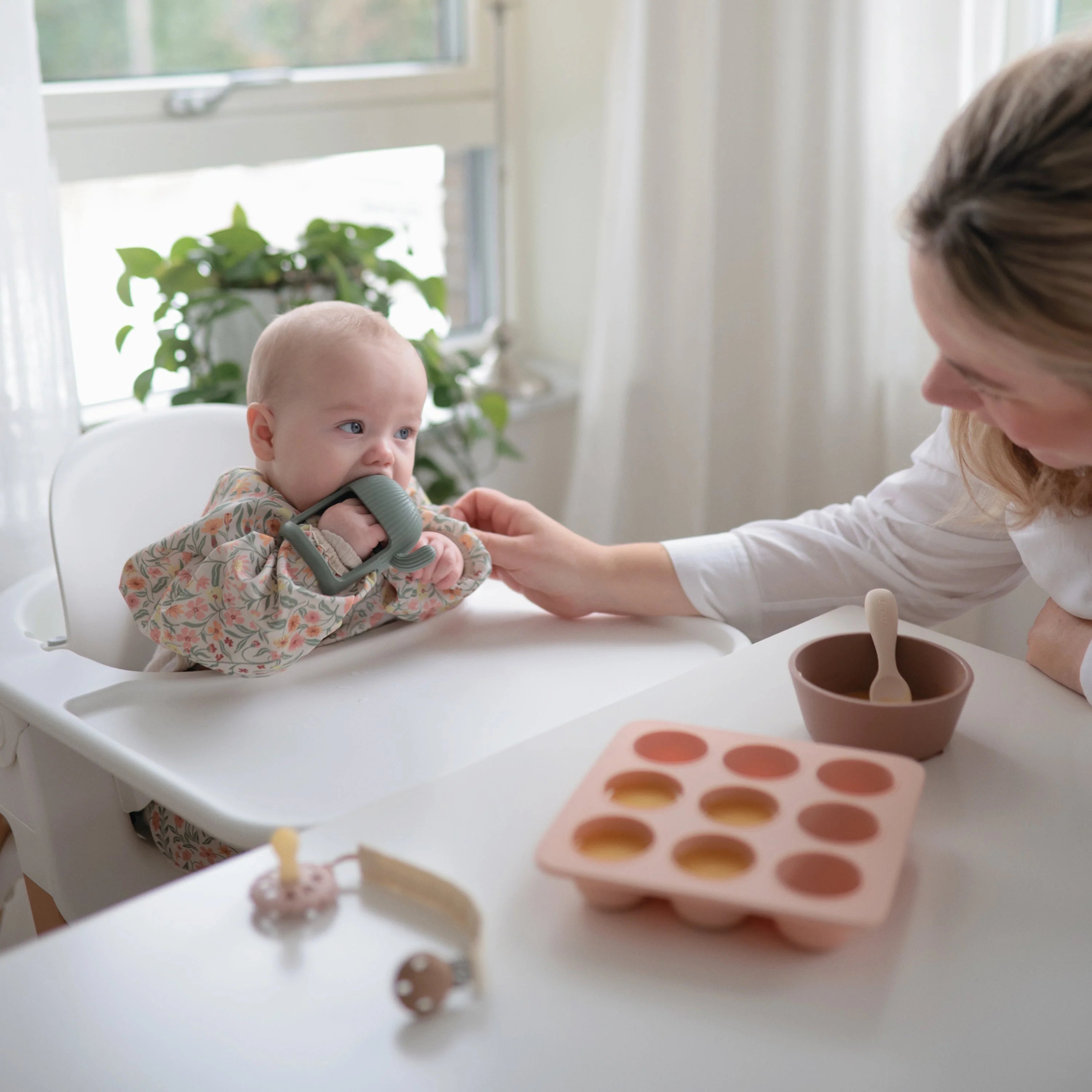 Baby in a high chair with the Mushie No-Drop Cactus Teether while a parent offers a snack
