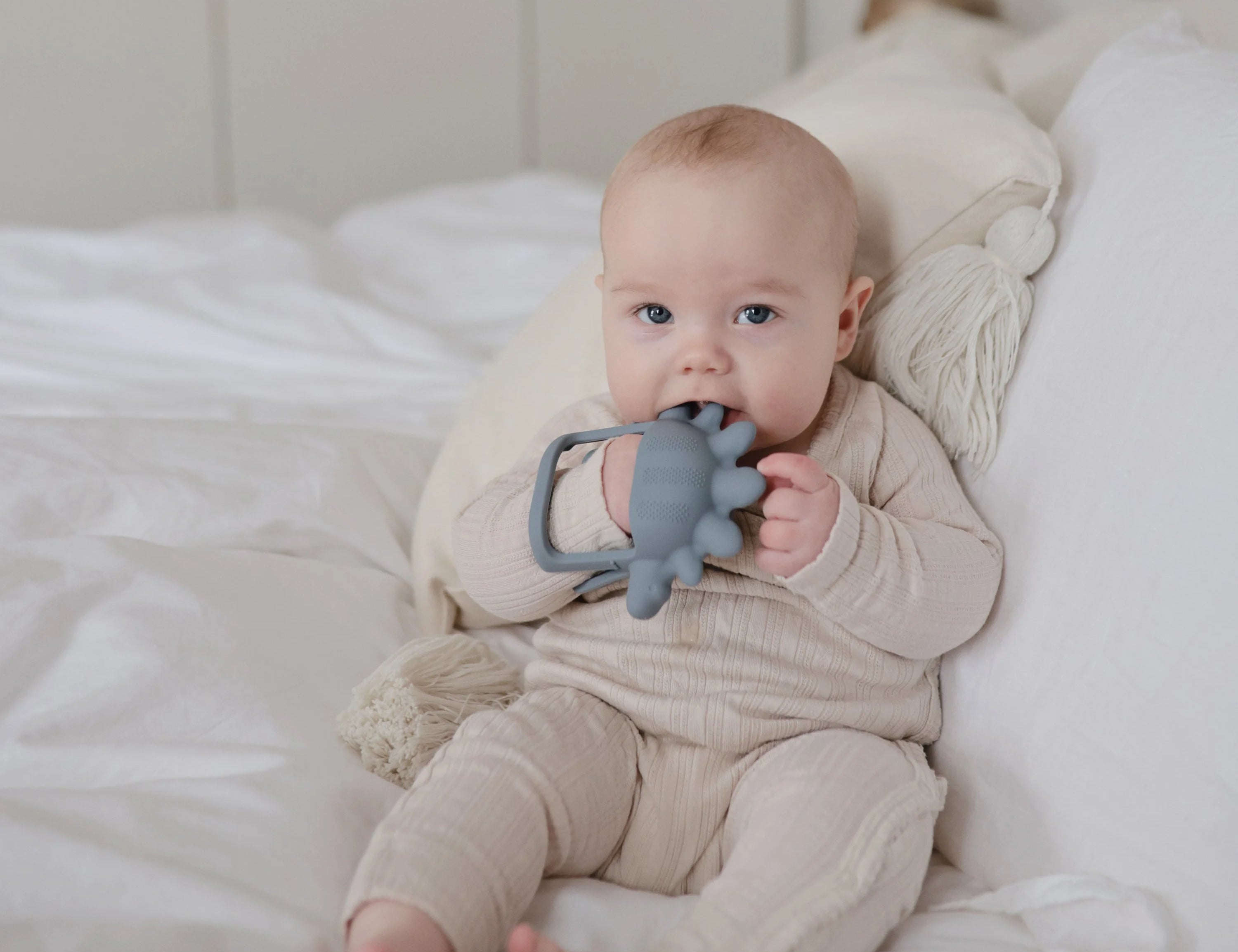 Baby lying on a bed chewing the Mushie No-Drop Dino Teether