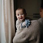 Smiling baby held by a parent holding the Mushie No-Drop Dino Teether