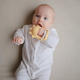 Baby in a white onesie lying down chewing the Mushie No-Drop Sun Teether