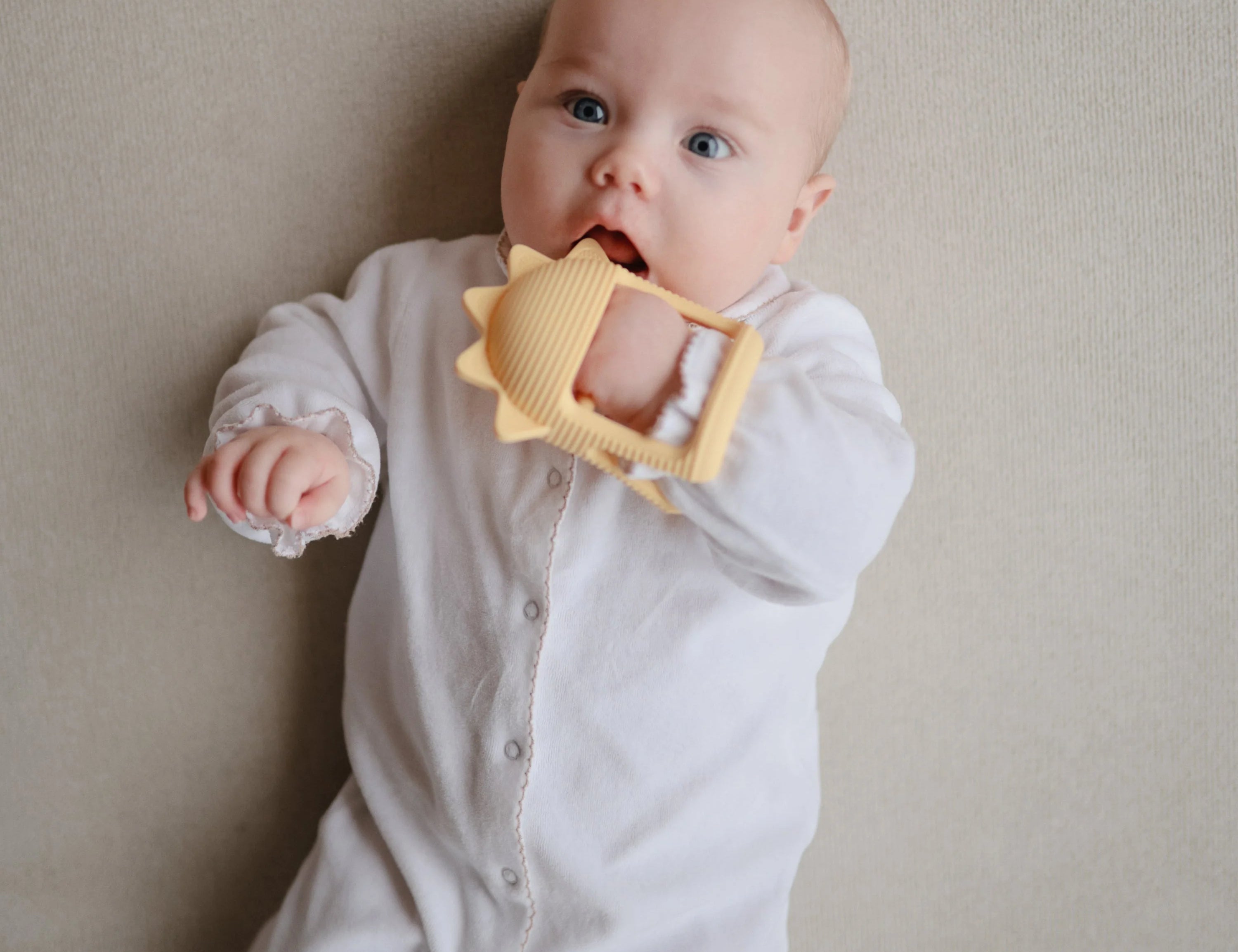 Baby in a white onesie lying down chewing the Mushie No-Drop Sun Teether