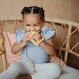 Laughing toddler in a rattan chair chewing the Mushie No-Drop Sun Teether