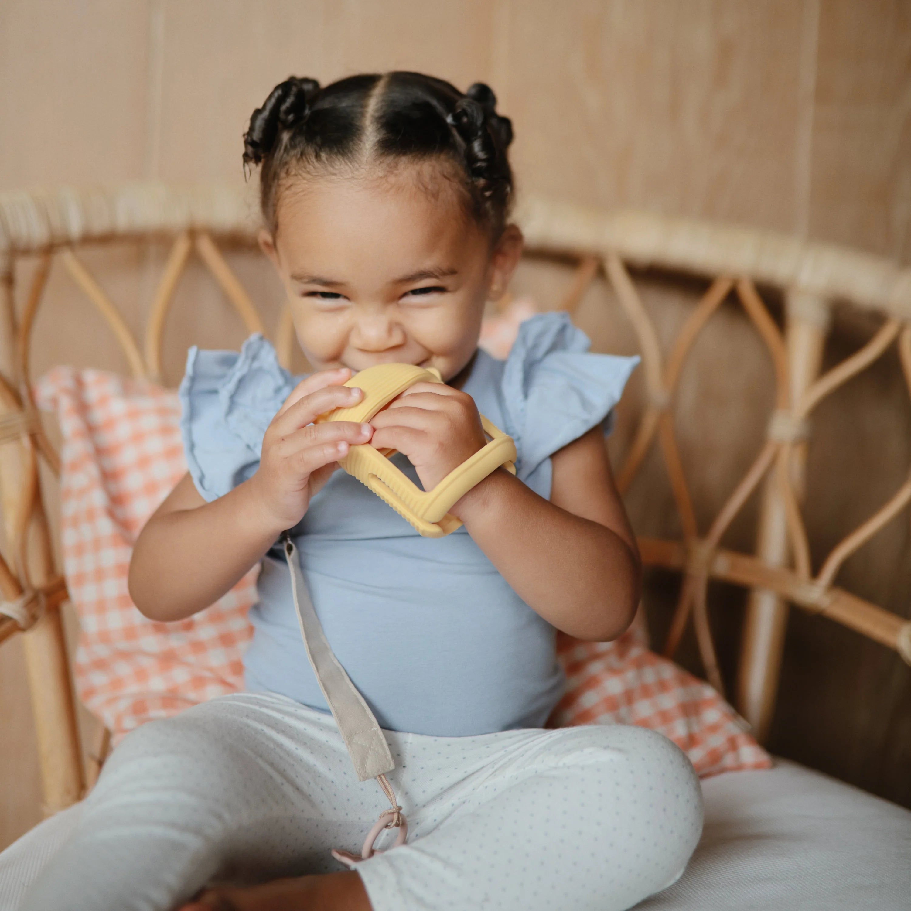Laughing toddler in a rattan chair chewing the Mushie No-Drop Sun Teether