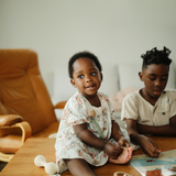 Two children sitting on a rug playing with Mushie toys in a living room
