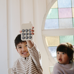 Smiling boy holding up the Mushie Phone Press Toy beside a younger child by a stained-glass window