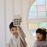 Smiling boy holding up the Mushie Phone Press Toy beside a younger child by a stained-glass window