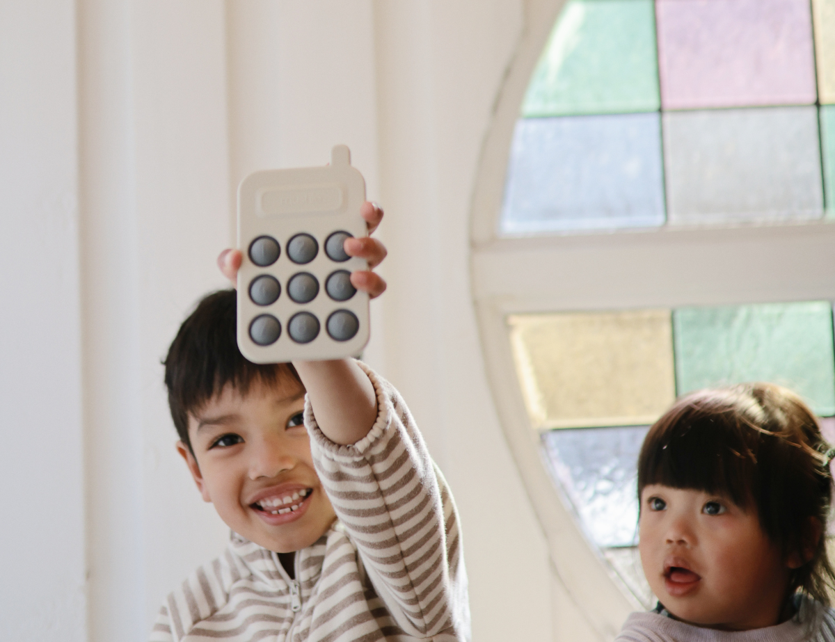 Smiling boy holding up the Mushie Phone Press Toy beside a younger child by a stained-glass window