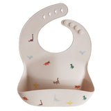 Mushie Silicone Baby Bib in Dinosaurs, a blush bib with a colorful dinosaur print and a catch pocket