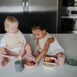 Two toddlers eating at a table wearing Mushie Silicone Baby Bibs